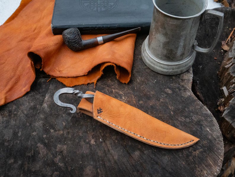 Pu&ograve; includere: Un coltello artigianale con una lama curva e un fodero in pelle. Il coltello &egrave; in metallo e ha un manico attorcigliato. Il fodero &egrave; marrone chiaro e cucito. Sono raffigurati anche una pipa, un boccale di peltro e una pelle di cuoio.