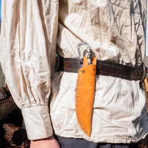 Pu&ograve; includere: Un fodero per coltello in pelle arancione caldo, cucito con un filo a contrasto. Il fodero &egrave; attaccato a una cintura in pelle marrone scuro con una fibbia in metallo. Un gancio in metallo forgiato &egrave; anch'esso attaccato alla cintura.