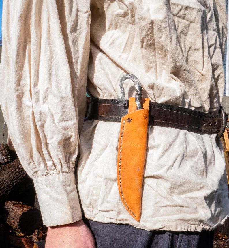 Pu&ograve; includere: Un fodero per coltello in pelle arancione caldo, cucito con un filo a contrasto. Il fodero &egrave; attaccato a una cintura in pelle marrone scuro con una fibbia in metallo. Un gancio in metallo forgiato &egrave; anch'esso attaccato alla cintura.