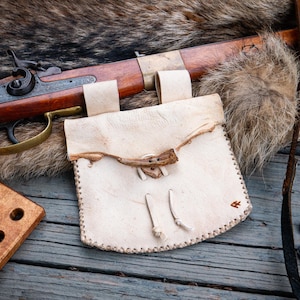 May include: A light-colored leather pouch with a flap secured by a small antler toggle, featuring stitched edges and two leather ties. The pouch is positioned near a wooden rifle with a brass trigger guard and a small wooden block with six holes.