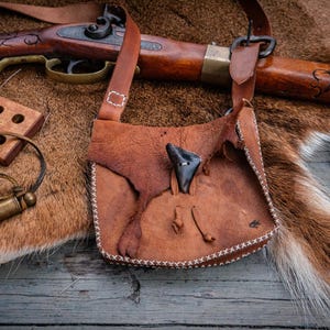 May include: A brown leather pouch with a flap closure secured by a dark triangular toggle, featuring visible white stitching along the edges. The pouch includes a long leather shoulder strap and is displayed alongside a wooden musket and accessories.