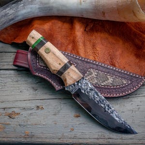 May include: A fixed-blade knife with a hammered steel finish, a light wood handle featuring dark wood and green accents, and a brass pin. The knife rests on a dark brown leather sheath with intricate stitching and a textured inlay pattern.