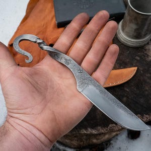 Pu&ograve; includere: Coltello in acciaio forgiato a mano con manico curvo e lama strutturata. Il coltello presenta un design a spirale sul manico e una punta affilata. La lama ha un motivo strutturato e la lettera "H" &egrave; stampata sulla lama.