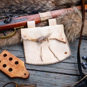 May include: A light-colored leather pouch with a toggle closure made from an antler piece, featuring stitched edges. Nearby are a wooden block with six circular holes, a long-barreled rifle with brass accents, and several small lead balls.