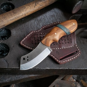 May include: A handcrafted knife with a wooden handle and a curved, silver blade. The knife rests on a brown leather sheath with a woven design. The handle has a teal accent and is secured with two small rivets.