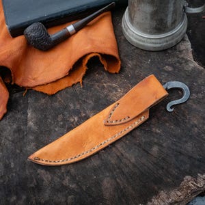 Pu&ograve; includere: Un fodero per coltello in pelle marrone chiaro, con un gancio in metallo curvo. Il fodero &egrave; cucito con un filo a contrasto. Una pipa e un libro sullo sfondo suggeriscono un tema rustico.