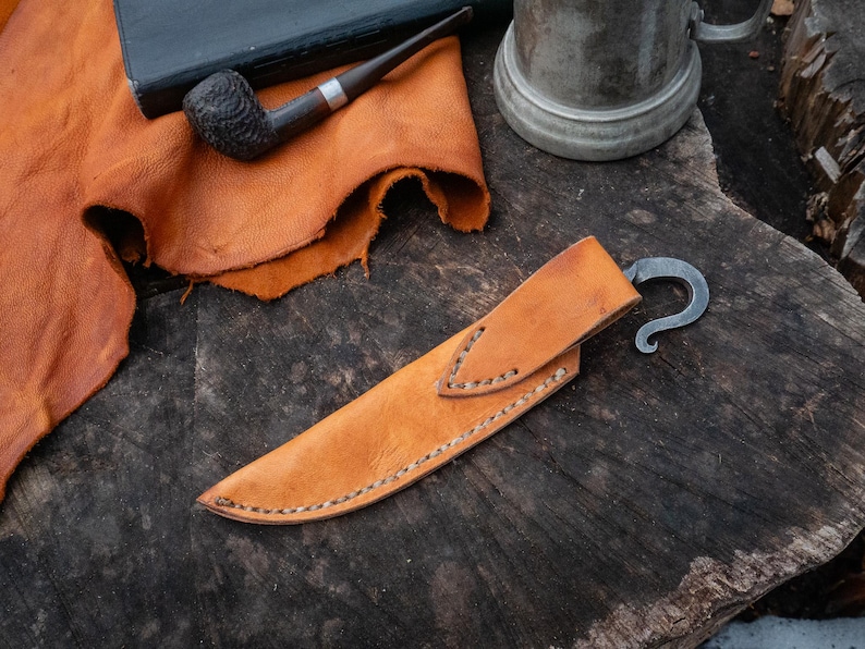 Pu&ograve; includere: Un fodero per coltello in pelle marrone chiaro, con un gancio in metallo curvo. Il fodero &egrave; cucito con un filo a contrasto. Una pipa e un libro sullo sfondo suggeriscono un tema rustico.