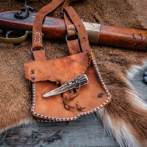 May include: A tan leather pouch with white stitched edges and a deer antler toggle closure. The pouch features a long leather strap and is positioned near a wooden musket with metal accents and a small pile of round lead musket balls.
