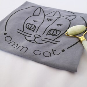 Puede incluir: Una camiseta gris con una ilustración de un gato negro y el texto "omm cat". Un rodillo de jade con mango dorado está sobre la camiseta.