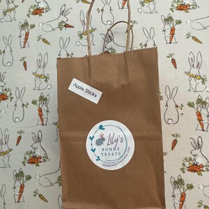 May include: A brown paper bag with a white label that says "Apple Sticks" and a white sticker with a bunny logo that says "Lily's Bunny Treats" and a website address. The bag is sitting on a white fabric with a pattern of bunnies holding carrots.