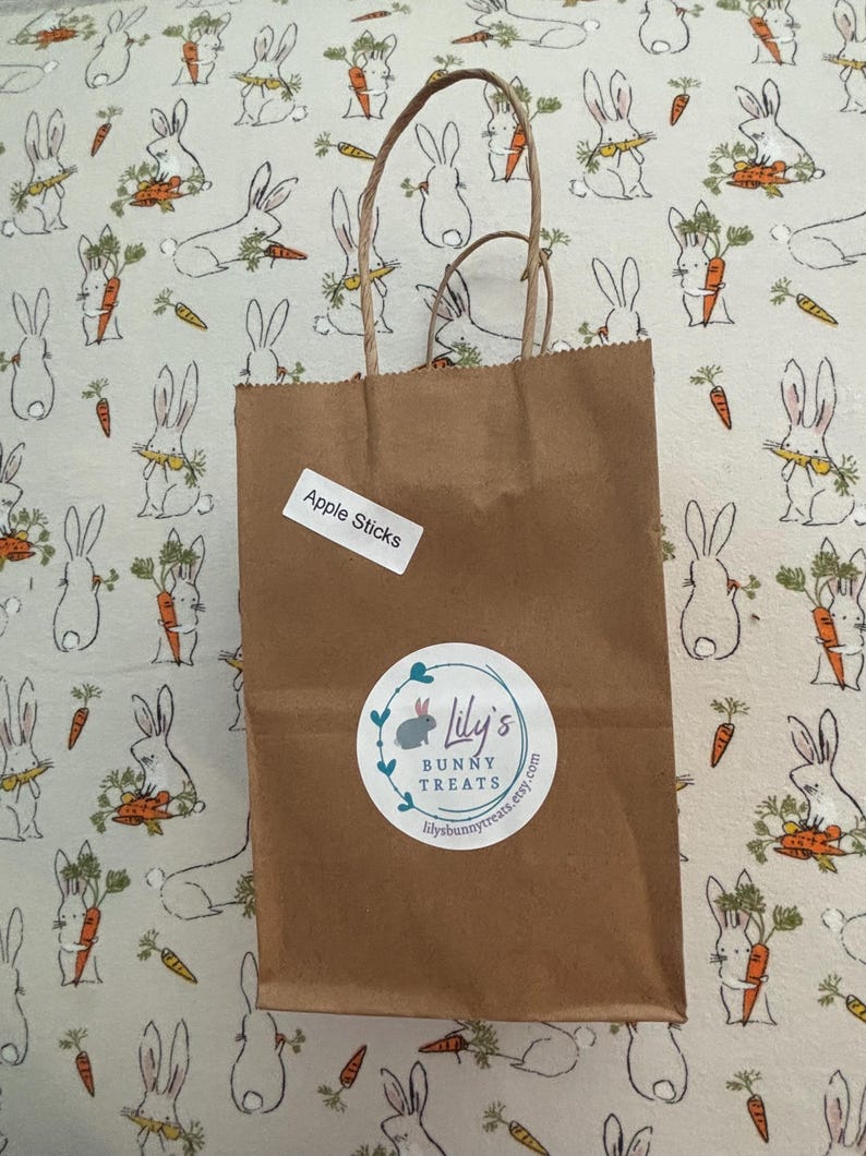 May include: A brown paper bag with a white label that says "Apple Sticks" and a white sticker with a bunny logo that says "Lily's Bunny Treats" and a website address. The bag is sitting on a white fabric with a pattern of bunnies holding carrots.