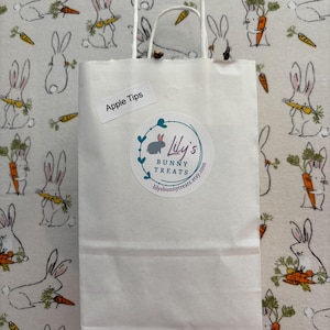 Puede incluir: Una bolsa de papel blanca con asa, etiquetada con "Apple Tips" y una pegatina que dice "Lily's Bunny Treats" con una direcci&oacute;n de sitio web. La bolsa est&aacute; sobre un fondo blanco con un patr&oacute;n de conejos blancos sosteniendo zanahorias.