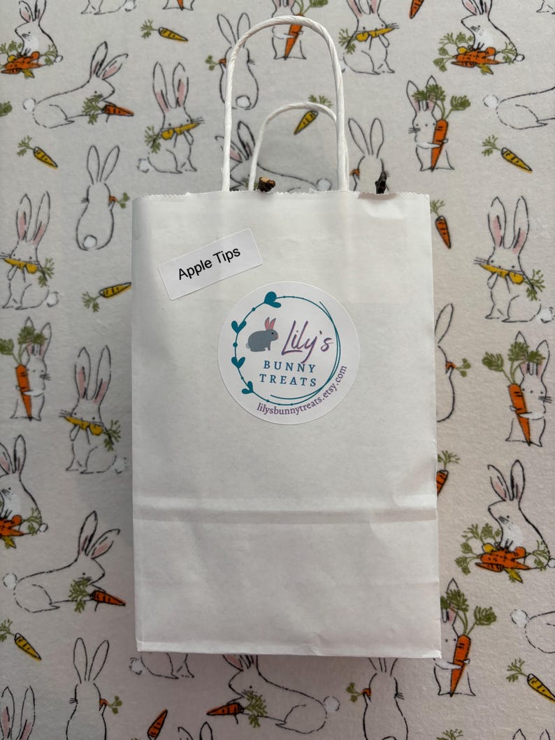 Puede incluir: Una bolsa de papel blanca con asa, etiquetada con "Apple Tips" y una pegatina que dice "Lily's Bunny Treats" con una direcci&oacute;n de sitio web. La bolsa est&aacute; sobre un fondo blanco con un patr&oacute;n de conejos blancos sosteniendo zanahorias.