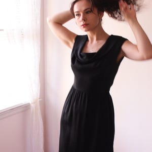 Może przedstawiać: Czarna sukienka z dekoltem typu łódka i krótkimi rękawami. Sukienka sięga do kolan i ma dopasowany gorset.