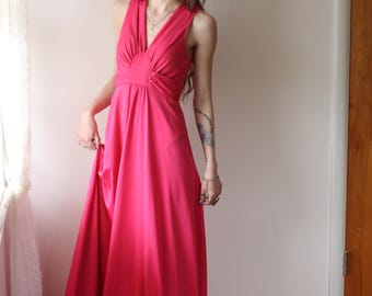 May include: A bright pink maxi dress with a halter neck and a flowing skirt. The dress has a gathered detail at the bust and is made of a smooth, solid fabric. The dress is floor-length and suitable for formal events.