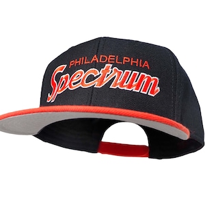 Op de afbeelding: Een zwarte en rode baseballpet met de tekst "PHILADELPHIA SPECTRUM" geborduurd in oranje en wit.