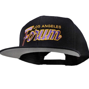 Può includere: Cappellino da baseball nero con visiera grigia. La parte anteriore del cappello presenta un logo ricamato in viola e giallo che dice "Los Angeles Forum".