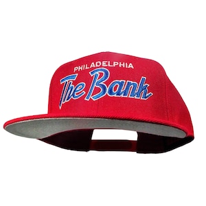 Op de afbeelding: Rode baseballpet met een grijze klep. De voorkant van de pet heeft de tekst "PHILADELPHIA The Bank" in blauwe en witte borduurwerk.