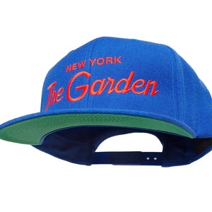 Op de afbeelding: Een blauwe en groene baseballpet met de tekst "NEW YORK The Garden" geborduurd in rood.