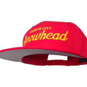 Puede incluir: Una gorra de béisbol roja y gris con el texto "Kansas City Arrowhead" bordado en amarillo.