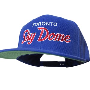 Op de afbeelding: Een koninklijk blauwe baseballpet met een groene onderkant. De voorkant van de pet heeft een witte geborduurde tekst met de tekst "Toronto Sky Dome" in een rood cursief schrift.