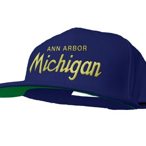 Op de afbeelding: Een marineblauwe baseballpet met een groene klep. De pet heeft de tekst "ANN ARBOR" en "Michigan" in goud geborduurd op de voorkant.