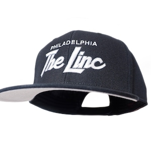 May include: A black and white baseball cap with the text "PHILADELPHIA The Linc" embroidered on the front.