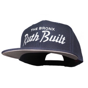 Puede incluir: Una gorra de béisbol azul marino con un logotipo bordado en blanco que dice "The Bronx Ruth Built".