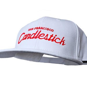 Op de afbeelding: Witte baseballpet met een rood geborduurd logo met de tekst "San Francisco Candlestick"