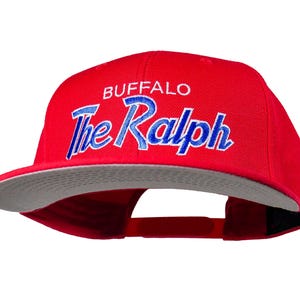 Puede incluir: Gorra de béisbol roja con visera plana y parte inferior gris. La gorra presenta la palabra "BUFFALO" en blanco y "The Ralph" en azul bordado en la parte delantera. Tiene una correa ajustable en la parte trasera.