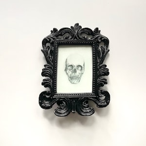 May include: A black ornate frame with a white mount and a detailed pencil drawing of a human skull.