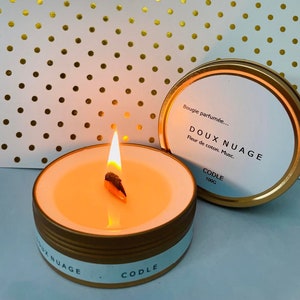 Peut inclure: Une boîte de bougie en or avec une étiquette blanche qui indique "Bougie parfumée... DOUX NUAGE Fleur de coton. Musc. CODLE 100G". La bougie est allumée et la cire est de couleur orange clair. La bougie est posée sur un fond blanc avec des pois dorés.