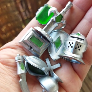 May include: A set of silver-colored miniature fantasy role-playing game accessories, including a sword, shield, helmet, and two other items. The accessories have green gem accents.