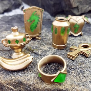 Könnte beinhalten: Ein Set aus goldfarbenen Miniaturfiguren mit grünen Akzenten. Das Set enthält einen Schild mit einem Drachenmotiv, eine Vase, einen Ring, einen Kelch, ein Bananenpaar und ein Ankh.