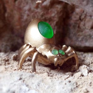 Könnte beinhalten: Eine goldfarbene Spinnenfigur mit grünen Edelsteinaugen und einem großen grünen Edelstein auf dem Rücken.