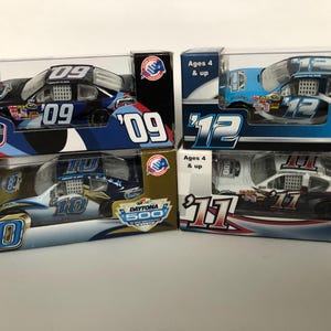 Puede incluir: Cuatro coches de carreras de juguete en sus cajas originales. Los coches son de varios colores, incluyendo negro, azul y blanco, con los números 09, 10, 12 y 11. Las cajas presentan el logotipo de Daytona 500.