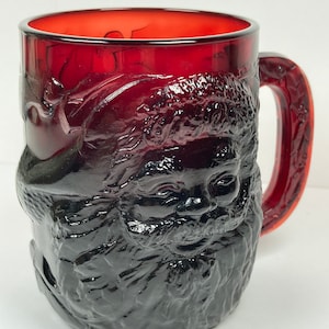 May include: A ruby red glass mug with a detailed embossed image of Santa Claus. The mug features a handle and a textured surface, perfect for holiday beverages. The Santa Claus design includes his face, beard, and hat.