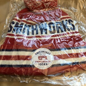 May include: Red and white striped knit beanie with a pom pom on top. The beanie has a blue and white patch with the text "Smithworks Trade Mark American Made Vodka" and a small tractor image.