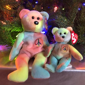 May include: Two tie-dye beanie baby bears, one large and one small, sitting in front of a Christmas tree. Both bears have a peace sign on their chest and are wearing Ty tags.