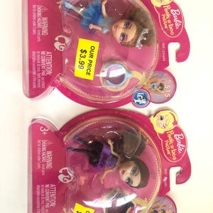 May include: Two Barbie Peek-a-boo Petites dolls in pink packaging. One doll wears a blue dress and the other a purple dress. The packaging includes a warning about small parts and the text "OUR PRICE $3.99".