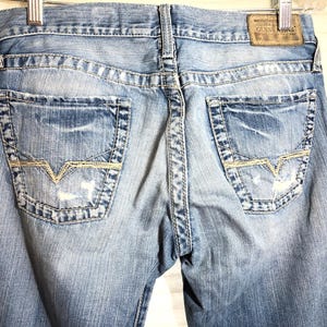 May include: Light blue denim jeans with distressed details, featuring back pockets with a decorative stitch and the Guess logo. The jeans have a faded wash and a leather patch.