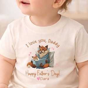 May include: Cream-colored t-shirt with a cartoon fox reading a book to a smaller fox. The text "I love you, Daddy" and "Happy Father's Day! Clara" are printed on the shirt. The design includes pink hearts.