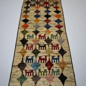 May include: A long, rectangular runner rug with a beige base featuring a repeating pattern of multicolored diamonds and stylized animal figures in red, blue, green, yellow, and brown. The rug has a dark brown border and short fringe at both ends.
