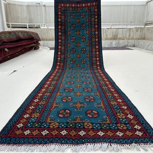 May include: A long, rectangular rug with a teal central field and intricate geometric patterns in red, brown, and white. The rug has a dark blue border and white fringe. The rug is displayed on a white surface.