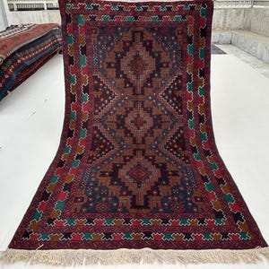 May include: A large, rectangular rug with a detailed geometric pattern in shades of brown, red, green, and blue. The rug has a central diamond design and a decorative border. The edges are finished with a fringe. The rug is displayed on a white surface.