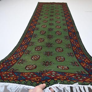 May include: Green runner rug with a repeating pattern of medallions and floral motifs. The rug has a border with geometric designs in red, blue, and orange. White fringe edges the bottom.
