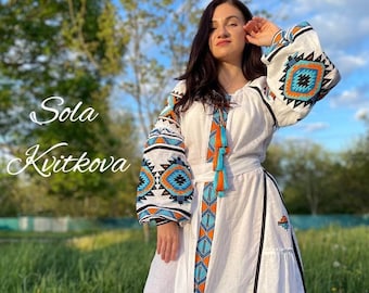 May include: A white linen dress with blue and orange geometric embroidery. The dress has a long skirt and a fitted bodice. The model is wearing the dress in a field of green grass.