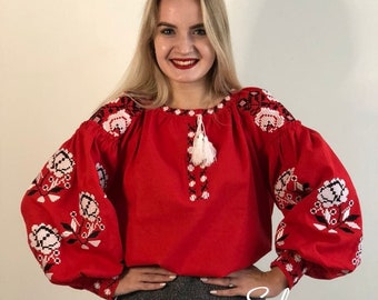 Könnte beinhalten: Eine rote bestickte Bluse mit weißen und schwarzen Blumendesigns. Die Bluse hat lange Ärmel und einen runden Ausschnitt. Das Model trägt einen grauen Rock und lächelt. Der Text "Sola Kvitkova" ist am unteren Rand des Bildes sichtbar.