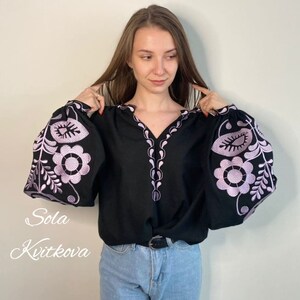 Op de afbeelding: Een zwart blouse met lange mouwen en een V-hals. De mouwen zijn versierd met paarse geborduurde bloemenmotieven. Het blouse wordt gedragen met een blauwe jeans.