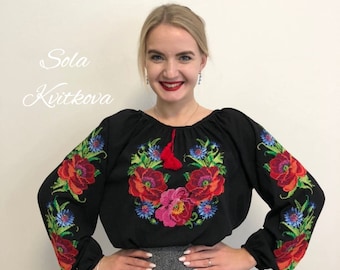 May include: A black embroidered blouse with a floral design in red, pink, blue, and green. The blouse has long sleeves and a round neckline. The embroidery features a wreath of flowers with a red tassel.